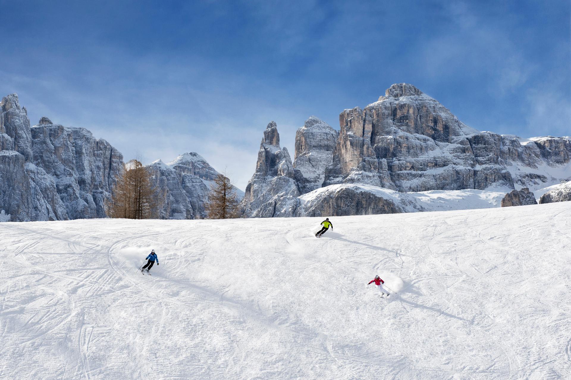 3 skiiers skiing down slope with rocky mountain peaks in background